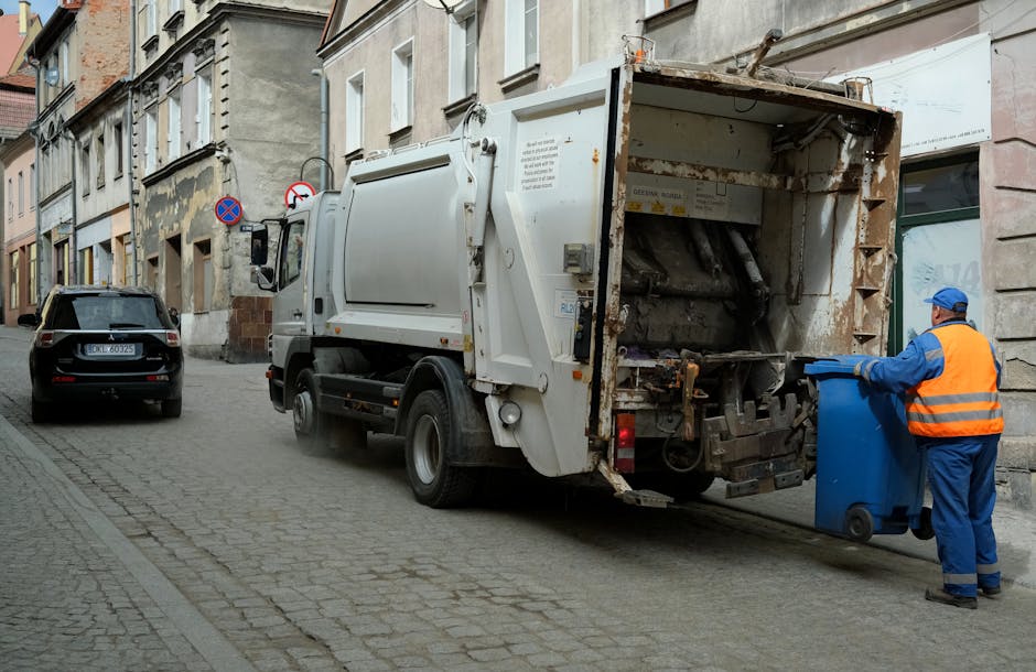 A street scene showing a white rubbish collection truck operated by Rubbish Removal Merton parked alongside a row of old buildings with weathered facades and peeling paint. The truck's rear compactor is open, revealing rusted metal panels, hydraulic mechanisms, and a worn, stained interior used for collecting waste. A worker dressed in a blue uniform and high-visibility orange vest is positioned on the right, emptying a blue wheelie bin into the truck’s hopper. The scene is illuminated by natural daylight, casting soft shadows on the cobbled pavement and street, which features a parked black car on the left and a no-parking sign attached to the building wall. The setting suggests an urban environment where private waste collection services, such as those provided by Rubbish Removal Merton, are used for on-site rubbish removal and disposal, contributing to alternative waste handling outside of local authority collection systems.