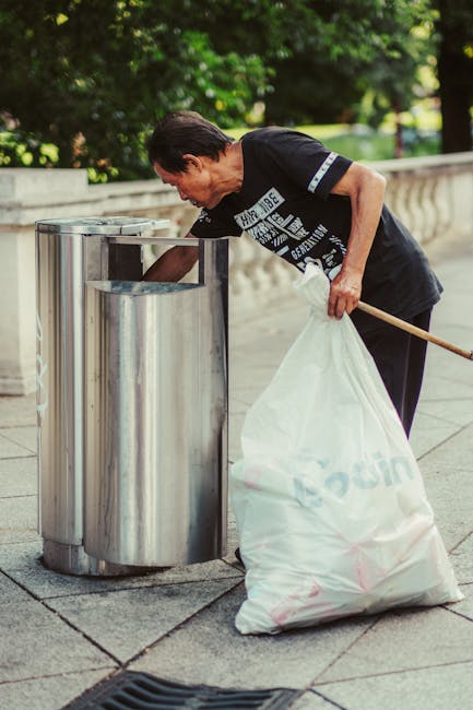 A middle-aged man with dark hair and wearing a black T-shirt is seen disposing of rubbish into a stainless steel public trash bin located on a paved sidewalk. The trash bin has three separate compartments designed for waste separation, each with a rectangular opening at the top. The man is holding a large white plastic bag full of waste, with the bag positioned on the ground beside him, and is leaning forward to deposit items into one of the compartments. The background features a lush, green outdoor setting with trimmed trees and shrubs, and a stone balustrade runs along the edge of the pavement. The scene is lit by natural daylight, creating a clear, detailed, and neutral representation of waste disposal activity, which aligns with the broader context of rubbish removal and waste management services such as those provided by Rubbish Removal Merton.