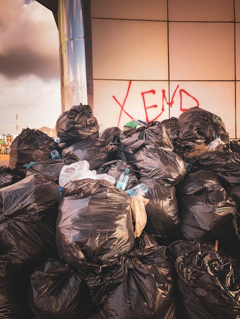 A large pile of black plastic rubbish bags stacked against a modern building with a tiled exterior wall. The bags are filled with various waste items, some slightly torn, revealing plastic bottles and packaging inside. The bags are arranged haphazardly, with a few white and transparent plastic bottles scattered among them, some partially visible and protruding from the bags. Behind the pile, there is graffiti with the word 'XEND' spray-painted in red on the wall. The scene is illuminated by natural light, casting soft shadows on the bags and emphasizing their glossy surface. The background shows a cloudy sky and some distant buildings, indicating an outdoor setting possibly near a commercial or urban area where independent waste disposal or rubbish removal services, such as those provided by Rubbish Removal Merton, are engaged for private on-site clearance or alternative rubbish collection.