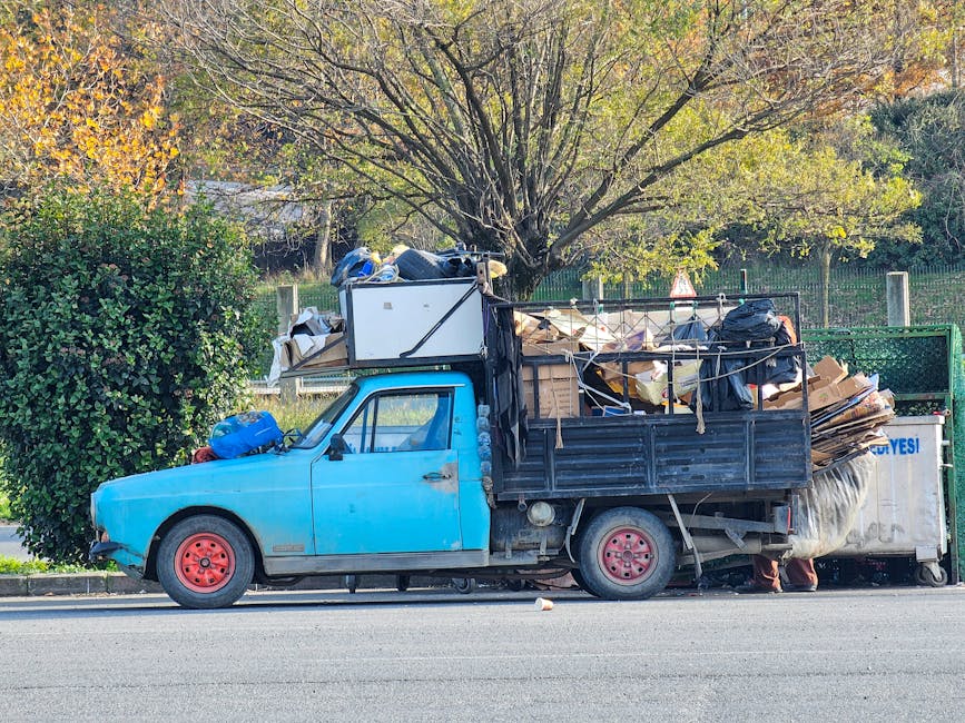 A small, light blue pickup truck with a black open cargo area parked beside a curb, loaded with various types of household waste and discarded materials including cardboard boxes, plastic bags, and miscellaneous debris. The vehicle's front wheels are painted red, and the truck shows signs of wear with some visible dirt and minor rust. The cargo area is densely packed and overfilled, with items extending beyond the sides and rear, secured loosely with ropes and strings. Behind the truck, there is a background of green trees with autumn foliage, including a large tree with thin branches and yellow-green leaves, and a wire fence running along the perimeter of a park or garden area. The scene is illuminated by natural daylight, creating a clear and neutral setting that suggests private, independent waste collection or on-site clearance, which Rubbish Removal Merton offers as part of its rubbish services in the local area.