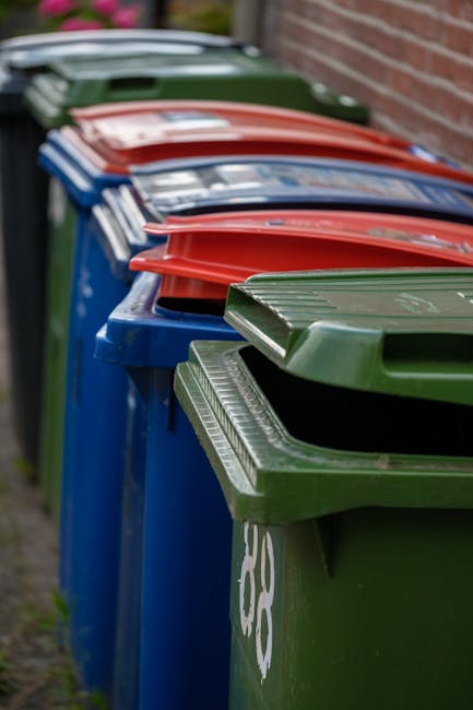 A worker wearing a red safety vest and black trousers is operating a large red rubbish collection vehicle on a paved street. The rear of the vehicle features a wide opening with mechanical arms and hydraulic components for lifting waste. The worker appears to be managing the waste materials, which include black plastic bags and assorted debris, some of which are on the ground near the vehicle. The debris consists of small items and loose rubbish, with a few scattered pieces on the pavement adjacent to a curb. To the right of the worker, there is a motorcycle parked on the pavement, with a small metal cage or barrier in front of it, possibly for additional waste collection tools or safety equipment. The surroundings include some greenery and trees in the background, along with a faint view of buildings and utility poles, indicating an urban or suburban environment. The scene is lit by natural daylight, with an overcast sky providing diffuse lighting, typical of outdoor waste management activities. The context suggests a private waste collection or on-site rubbish removal service, consistent with independent rubbish disposal operations like those offered by Rubbish Removal Merton.