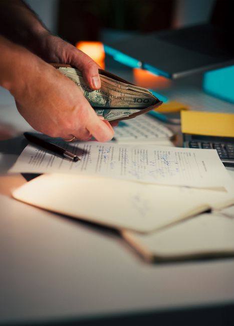 A close-up of a person's hand holding a fan of various banknotes, predominantly £100 notes, above a cluttered office desk. The desk features an open stack of papers with handwritten notes, a pen lying across some documents, and a calculator. In the background, there is a computer keyboard and a blurred out monitor, suggesting an indoor office setting. The lighting is soft, with a warm glow on the hand and cash, highlighting the textures of the paper currency and the smooth surface of the documents. The scene conveys themes related to financial management, budgeting, or business transactions, aligning with the context of independent collection or private waste disposal services provided by Rubbish Removal Merton, subtly hinting at the importance of cost considerations in rubbish contracts and alternative waste handling options in the Merton area.