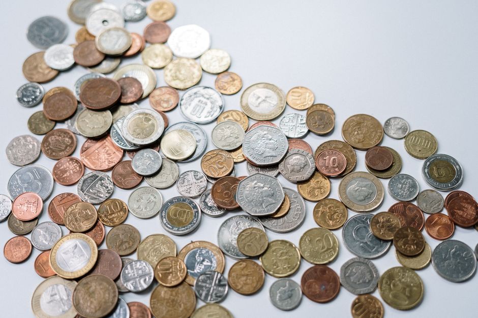 A scattered collection of various UK coins is displayed on a plain, light-colored surface. The coins include a mix of copper, silver, and bi-metallic types, with denominations ranging from 1p and 2p to higher values such as 10p, 20p, 50p, and £1 coins. The coins are arranged in a loose, overlapping pattern with some coins laying flat, showing both obverse and reverse designs, and others tilted or partially stacked. The surface appears smooth and clean, with no other objects visible in the immediate environment. This image, though primarily of coins, subtly relates to the concept of costs associated with independent waste disposal or private rubbish collection services, such as those provided by Rubbish Removal Merton, by highlighting the potential expenses involved in alternative rubbish management options outside of municipal disposal channels.