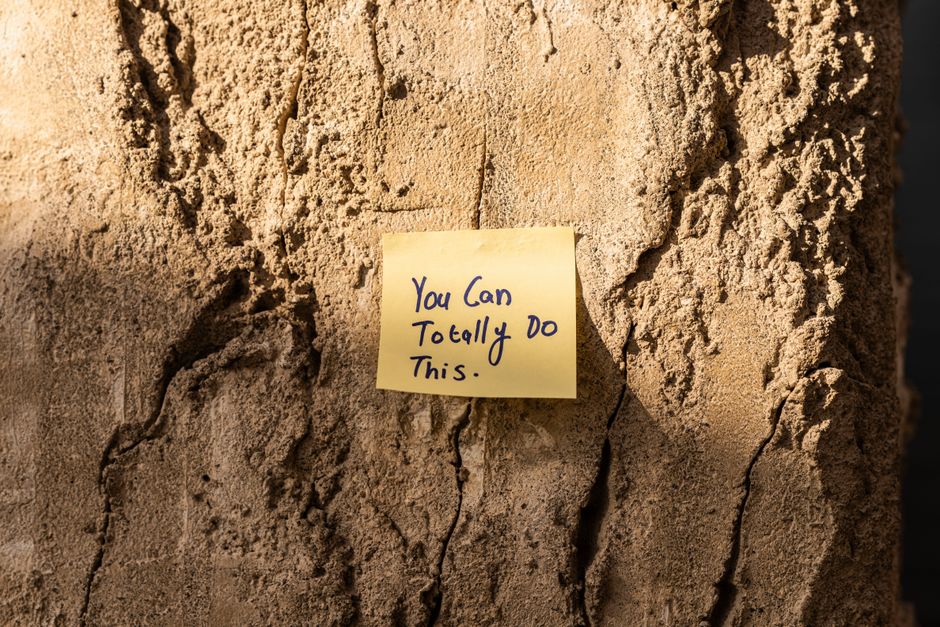 A close-up image of a textured, rough surface of a tree trunk with visible vertical fissures and bark layers in shades of light brown and tan. Affixed to the trunk is a small, yellow sticky note with handwritten text in blue ink that reads, 'You Can Totally Do This.' The note is positioned slightly off-center to the right and is adhered flat against the bark, with some shadowing around its edges. The background is blurred, emphasizing the detail of the tree bark and the motivational note, creating a natural outdoor setting that subtly relates to the theme of overcoming challenges. This scene may evoke themes of persistence and encouragement within outdoor or natural environments, aligned with the idea of taking on tasks such as on-site waste management or independent disposal efforts. The lighting is soft, highlighting the texture of the bark and the note without harsh shadows or glare, maintaining a neutral and professional tone that supports accessibility and contextual relevance to waste management and rubbish removal services.