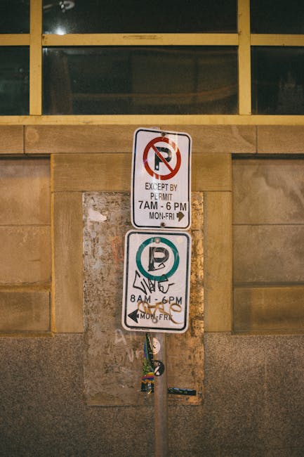 A vertical metal pole with two rectangular parking restriction signs attached, positioned in front of a textured stone wall with a large window above. The top sign indicates no parking except by permit from 7 am to 6 pm, Monday to Friday, with a red circle and slash over a black 'P' on a white background, and the text in black. The lower sign shows designated parking hours from 8 am to 6 pm, Monday to Friday, featuring a blue circle with a white 'P' symbol and an anti-clockwise arrow, accompanied by additional black text. The pole has some attached multi-colored stickers near its base. The background includes a section of the building's stone façade and large glass window panels framed with yellowish metal at the top, suggesting an urban environment potentially related to private property access or restricted parking zones. Rubbish removal companies like Rubbish Removal Merton may deal with waste located in such areas, offering solutions related to alternative disposal methods or clearance, especially where parking restrictions might influence logistics and collection efforts.
