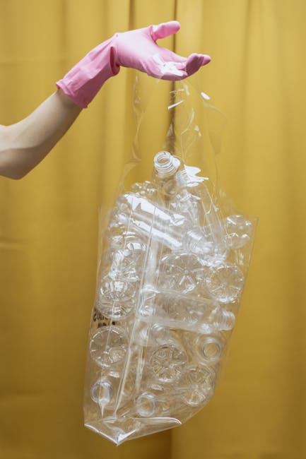 A person wearing a pink rubber glove is holding a clear plastic bag filled with crumpled and whole transparent plastic bottles and containers against a yellow background. The plastic items are of various shapes and sizes, primarily lightweight and used beverage bottles, some with visible necks and caps, and others with indentations or ridges. The bag appears partially filled, with the bottles inside loosely arranged, and the person's hand is gripping the top of the bag, which is open at the neck. The scene suggests handling or preparing plastic waste for disposal or recycling, consistent with rubbish removal or waste management activities. The yellow background provides a high-contrast setting, emphasizing the transparency of the plastic materials and the bright pink color of the glove used for handling the waste, supporting a professional and neutral observation relevant to waste collection services.