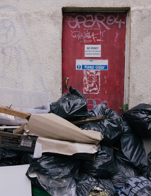 A collection of black rubbish bags, various cardboard pieces, and discarded household waste piled against a weathered red metal door mounted on an exterior wall of a building. The door features graffiti tags in white and pink spray paint, along with a white and blue sign that reads 