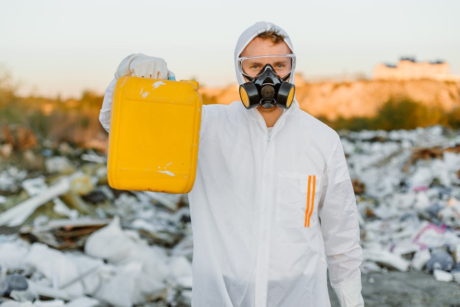 A person dressed in a white protective suit, including a hood and orange safety stripes, is standing outdoors in a rubbish-filled environment, holding a bright yellow plastic container with a slightly worn surface in their right hand. The individual is wearing a black and yellow respirator mask and clear safety goggles, suggesting involvement in hazardous waste handling or contamination control. The background reveals a terrain cluttered with scattered waste materials, likely consisting of plastic, paper, and other debris, with a blurred landscape in the distance under soft, natural lighting. The scene indicates an on-site waste clearance or rubbish removal operation, possibly related to hazardous waste laws or private disposal services. The overall environment emphasizes the importance of professional waste management, as conducted by companies such as Rubbish Removal Merton, in managing environmentally sensitive waste in compliance with legal standards.