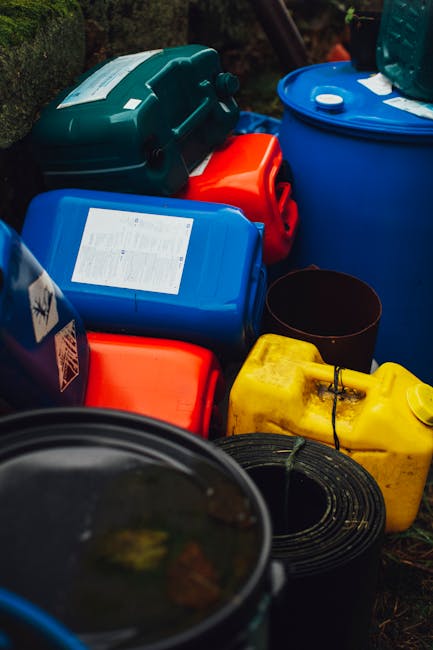 The image displays a variety of plastic containers and jerry cans arranged on a gravel or dirt surface, with several stacked or positioned side by side. The containers are made of durable plastic and come in colours including blue, red, yellow, and black, with some featuring labels or stickers indicating contents or handling instructions. A large blue barrel with a lid is partially visible on the right side, while smaller jerry cans and rectangular containers are in the foreground and background, some with spouts or handles for pouring. The textures range from smooth, glossy surfaces on the plastic to the rough, natural appearance of the outdoor environment. The scene appears to be outdoors, possibly in a garden or yard, amidst greenery or fencing, and the lighting suggests natural daylight. This arrangement of plastic waste containers suggests an on-site collection or storage area, typical of facilities or services involved in private waste handling or alternative rubbish collection, as managed by professional waste removal firms like Rubbish Removal Merton, aiming to comply with hazardous waste laws and safely store or prepare waste for proper disposal.