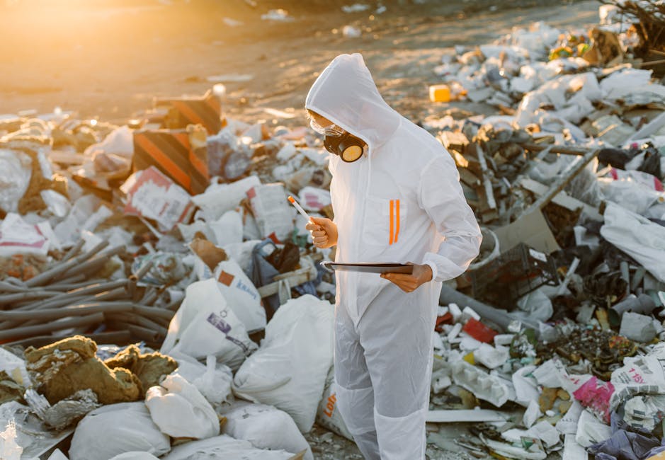 A person dressed in a white protective suit, including a hood and orange safety stripes, is standing outdoors in a rubbish-filled environment, holding a bright yellow plastic container with a slightly worn surface in their right hand. The individual is wearing a black and yellow respirator mask and clear safety goggles, suggesting involvement in hazardous waste handling or contamination control. The background reveals a terrain cluttered with scattered waste materials, likely consisting of plastic, paper, and other debris, with a blurred landscape in the distance under soft, natural lighting. The scene indicates an on-site waste clearance or rubbish removal operation, possibly related to hazardous waste laws or private disposal services. The overall environment emphasizes the importance of professional waste management, as conducted by companies such as Rubbish Removal Merton, in managing environmentally sensitive waste in compliance with legal standards.