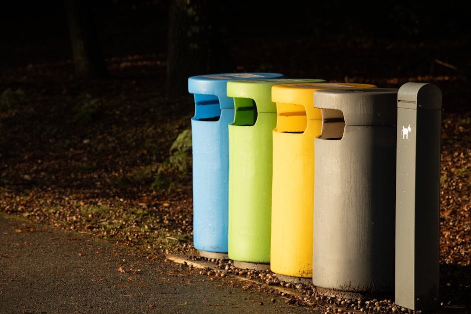 A row of four cylindrical public waste bins with brightly coloured plastic lids, positioned along the edge of a paved pathway in an outdoor setting during daylight. The first bin from the left has a blue lid, the second features a green lid, the third has a yellow lid, and the fourth is topped with a beige or light brown lid. Each bin appears to be made of weather-resistant plastic with a smooth, matte finish, and they are standing on a gravel bed next to the pathway. To the right of this row, there is a narrow, tall gray metal recycling or waste collection bin with a small pet waste icon printed on it. The background shows a natural environment with blurred foliage and trees, indicating an outdoor park or communal area. The scene depicts a typical setup for community rubbish disposal, possibly supporting private or independent waste collection services such as those offered by Rubbish Removal Merton, highlighting the importance of accessible waste separation points.