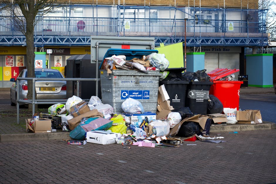 A large pile of mixed household rubbish and packaging waste accumulated around a public recycling bin station on a paved urban street. The waste includes cardboard boxes, plastic bags filled with various items, paper, and miscellaneous packaging materials, some of which are spilling onto the surrounding pavement. The recycling bins, which are identifiable by their labels and color codes, are positioned in front of a building with storefronts and a metal scaffolding structure on the upper level. A silver car is parked nearby, partially obscured by a metal railing and a small tree with a bare trunk. The scene appears to be outdoors during an overcast day, with diffused lighting highlighting the disorganized placement of waste, which suggests improper disposal or an overflow situation. Occasional signs on the building suggest this area may be associated with municipal waste collection points, with some context related to independent rubbish removal or private waste handling services like Rubbish Removal Merton.