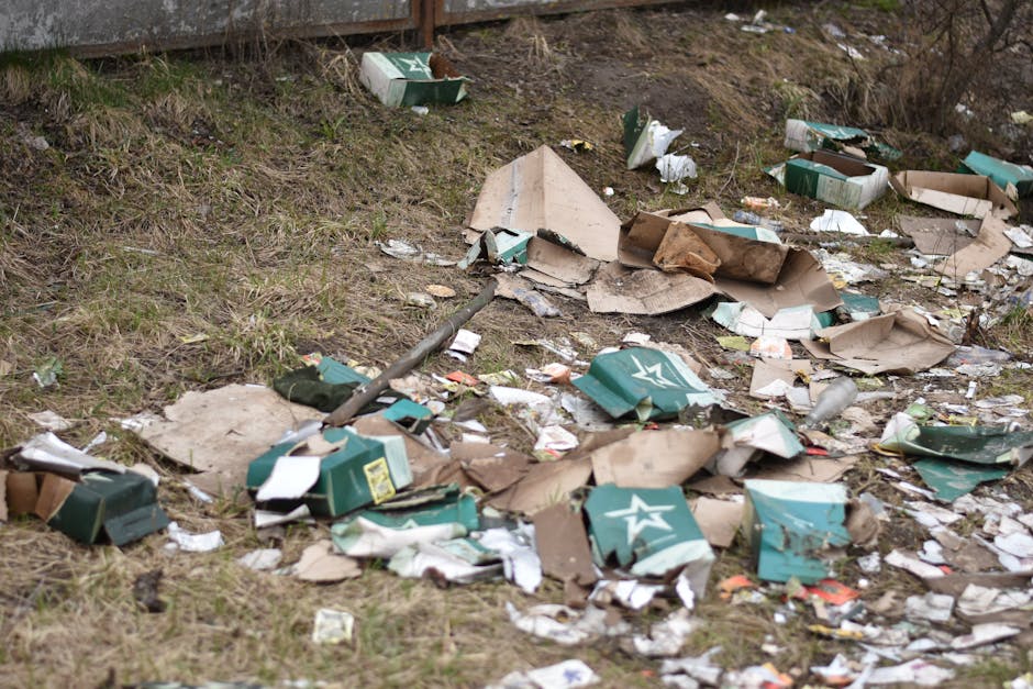 A patch of ground near a fence shows scattered debris including crumpled cardboard boxes, some of which are green with white logos, and torn paper. The boxes are partially flattened and mixed with smaller waste items and paper scraps, all lying on a grassy surface with patches of dirt. A metal pole or rod is visible among the rubbish, leaning on the ground and partially covered by the debris. The background features a wooden fence with some soil and sparse vegetation behind it, indicating an outdoor environment that may be a neglected or unmanaged area. The scene reflects typical roadside or park litter, highlighting the importance of rubbish removal services like those offered by Rubbish Removal Merton for effective waste disposal and environmental cleanup.