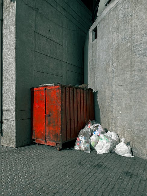 A large, industrial-style red metal skip with visible rust and chipped paint, positioned between a dark green wall with horizontal panels and a light grey concrete wall next to a building's exterior. The skip is partially full, with several clear plastic rubbish bags filled with assorted waste materials stacked beside it on the paved ground. The bags contain mixed refuse, including plastic bottles and household rubbish. The paving surface consists of dark grey, textured concrete tiles, and the scene is illuminated by natural daylight, casting minimal shadows. The setting appears to be an urban alley or service area, reflecting a typical scene where private waste collection or rubbish removal services, like those offered by Rubbish Removal Merton, could facilitate the clearance of unmanaged waste in a designated outdoor space near commercial or residential buildings.