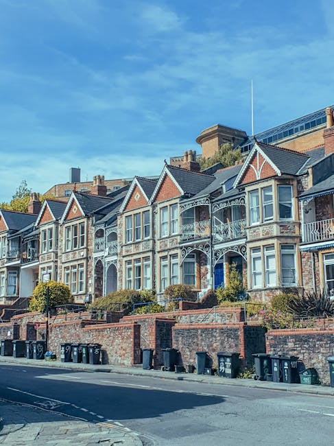 A row of Victorian-style terraced houses with ornate white ironwork balconies and decorative trim, positioned behind a brick retaining wall with small shrubs and bushes in front. The houses feature bay windows with white frames, grey slate roofs, and brick walls in shades of red and brown. In the foreground, a paved street with multiple black wheelie bins aligned along the curb is visible, suggesting waste collection activity. The scene is illuminated by natural daylight under a clear blue sky, with neighboring modern high-rise buildings in the background, indicating an urban residential area. This setting reflects typical private waste management practices with on-site rubbish storage, aligning with the context of independent rubbish removal services in Merton. The presence of waste bins adjacent to the properties underscores the importance of efficient rubbish disposal, consistent with professional rubbish removal solutions offered by Rubbish Removal Merton.