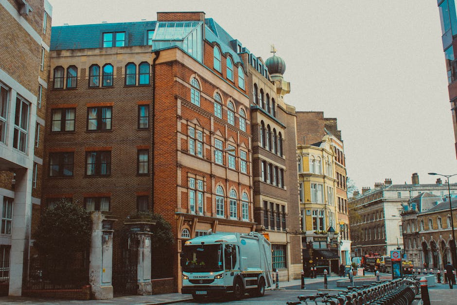 A city street scene featuring a mix of historic and modern brick buildings with multiple floors and ornate window designs. The foreground shows a light blue waste collection truck with a closed top, parked adjacent to a sidewalk. Surrounding the truck are small bollards and a row of parked bicycles. To the left, part of a white modern building with large windows is visible, while further down the street there are additional buildings with varying architectural styles, including one with a yellow facade and intricate detailing. The street has minimal traffic, with a few other vehicles in the distance, and the sky overhead appears overcast, casting diffuse natural light across the scene. This urban environment, with its mix of architectural elements and the presence of waste collection, subtly relates to private rubbish removal and on-site disposal services, as handled by providers like Rubbish Removal Merton.