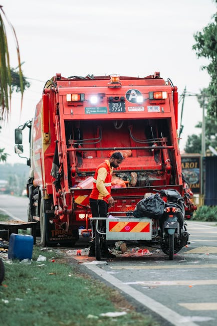 A sanitation worker wearing a yellow and red uniform is operating a red rubbish collection truck on a roadside during daytime. The worker is overseeing the emptying of waste into the truck's rear, which has an open hatch showing mechanical components and a cleaning mechanism. The truck is parked partly on the paved road adjacent to a grassy verge with a blue plastic container and a black motorbike positioned nearby. Behind the truck, there are trees, utility poles, and a building with signage, indicating an urban or suburban environment. The scene is lit by natural daylight, with a cloudy sky overhead, highlighting the materials of the waste bin, the texture of the truck’s metallic surface, and the work being carried out. Such scenes are typical of on-site waste collection and private rubbish removal services that may be part of professional rubbish management solutions in the area, as offered by Rubbish Removal Merton.