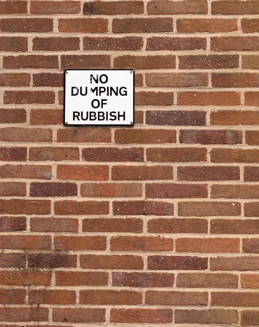 A rectangular white metal sign with black text reading 'NO DUMPING OF RUBBISH' is mounted on a red brick wall with a traditional running bond pattern. The bricks vary in shades of red and brown, with some darker and weathered, and are finished with light gray mortar. The sign is positioned slightly above the center of the image, with the wall extending horizontally in both directions. The lighting is even, highlighting the texture of the brick surface. This scene suggests a location where waste disposal or rubbish removal is regulated, and the signage indicates regulations related to private rubbish collection or current waste management policies. The presence of the sign in this setting reflects efforts by local waste removal specialists, such as Rubbish Removal Merton, to promote responsible waste handling and prevent illegal dumping in the community.