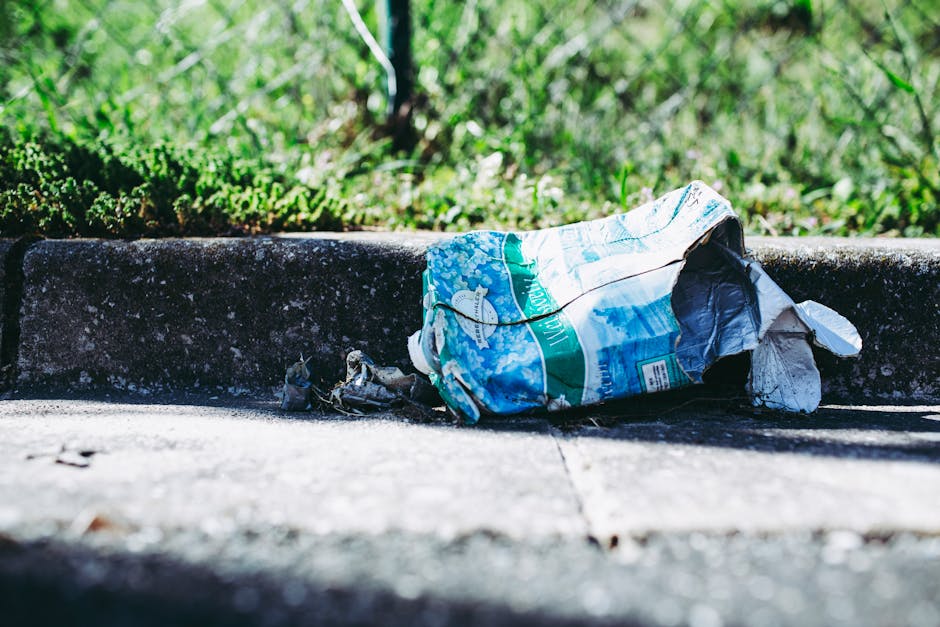 A crumpled plastic bag with a predominantly blue and white design is lying on the pavement near the curb, partially pressed against the raised edge of the sidewalk. The bag appears to contain trash and is coated in dust, with some torn sections exposing its contents. The surrounding area includes a concrete curb with a rough, weathered surface, and the pavement shows minor cracks and dirt. In the background, there is a grassy verge with dense, green vegetation, slightly out of focus, indicating an outdoor urban or suburban environment. The lighting suggests natural daylight, casting subtle shadows around the litter. The scene reflects an instance of improperly disposed waste, which could be targeted by rubbish removal services. Rubbish Removal Merton may handle such debris as part of their private waste clearance solutions, offering an alternative to on-site or municipal rubbish collection, with focus on maintaining cleanliness in public and private areas.